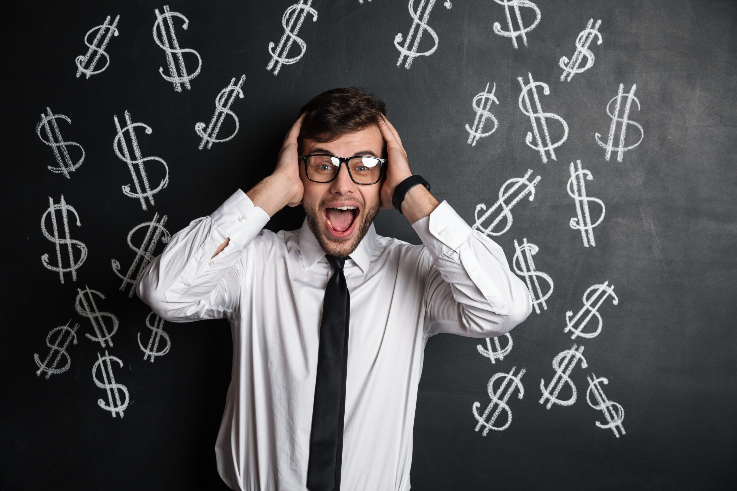 Overjoyed young bearded man in glasses holding his head, looking at camera, isolated over  blackboard with dollars symbol